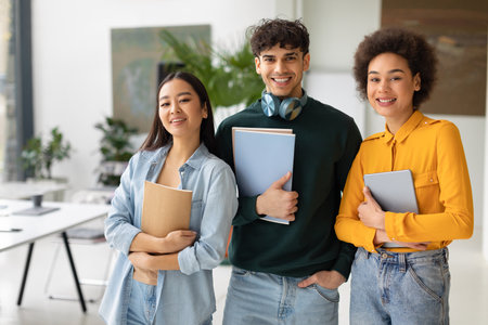 Happy three multiracial students posing in coworking space, young people with workbooks standing in classroomの写真素材
