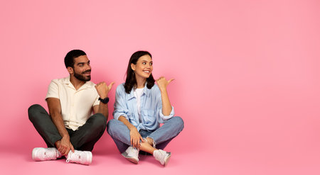 Curious happy couple sitting on floor, pointing at copy spaceの写真素材