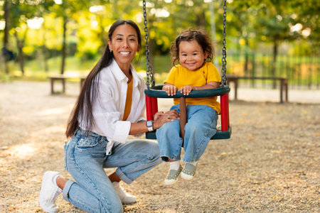 Cheerful Chinese Mom Riding Infant Daughter On Swings At Playground