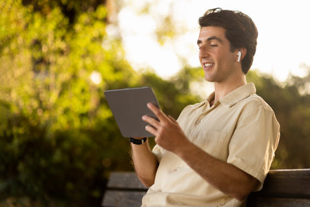 Cheerful man enjoying nice podcast, resting on bench at parkの写真素材