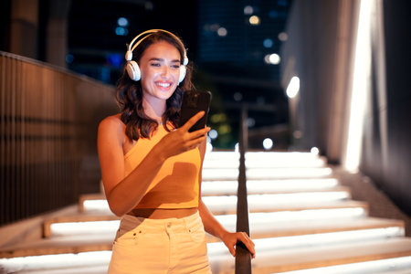 Beautiful trendy young european woman in headphones with smartphone listening music, walking in city in the eveningの写真素材