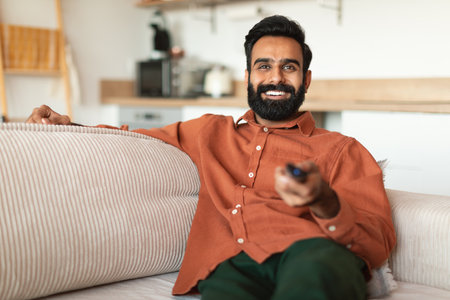 Cheerful Indian bearded man watching television show switching channels indoorsの写真素材