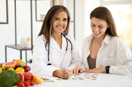 Smiling mature caucasian doctor nutritionist in white coat consultation young lady at table with organic vegetablesの写真素材