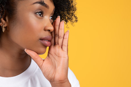 Cheerful young black curly lady in white t-shirt puts her hand to her face whispers