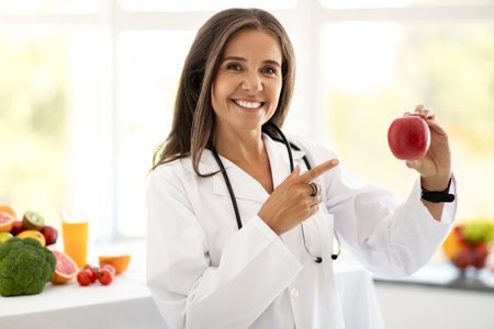 Smiling mature caucasian lady doctor nutritionist in white coat show appleの写真素材