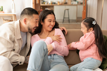 Asian family laughs together as daughter tickles parents at homeの写真素材