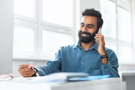 Smiling indian businessman talking on cellphone and noting down information, sitting at desk in officeの写真素材