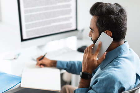 Indian businessman having conversation on smartphone, sitting in front of computer monitor, taking notes, rear viewの写真素材