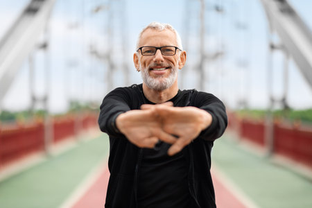 Happy well-fit senior sportsman exercising on city bridge, stretching handsの写真素材