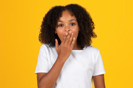 Positive shocked teenage black pupil girl in white t-shirt covers mouth with handの写真素材