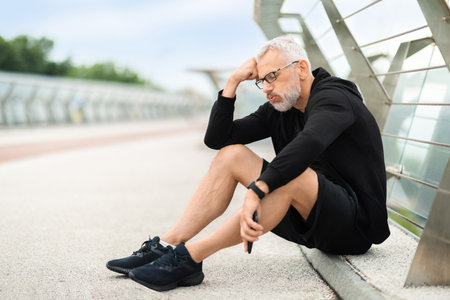 Exhausted elderly sportsman sitting on ground, working out outdoorの写真素材