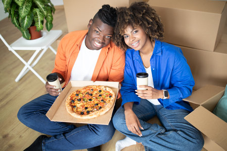 Cheerful black spouses eating pizza and drinking takeaway cofee in new homeの写真素材