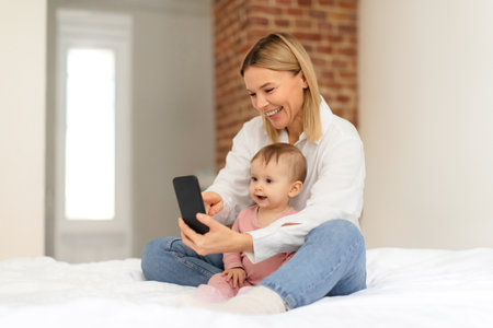 Cheerful mom and her infant child daughter using cellphone, surfing web for educational mobile games, sitting on bedの写真素材