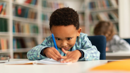 Concentrated black schoolboy sitting at desk in school and learning to write in copybook with pencil, studying, panoramaの写真素材