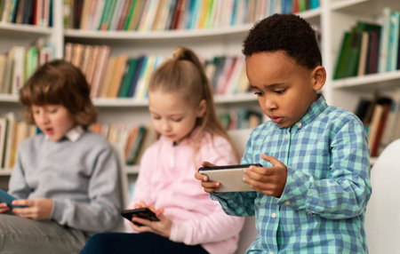 Diverse school kids students using smartphones in library, multiracial children holding mobile phones, playing gamesの写真素材