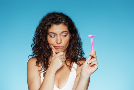 Pensive young european curly woman thinking and looking at razor in hand, isolated on blue backgroundの写真素材