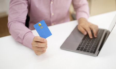 Caucasian mature guy at table with laptop uses credit card in office interior. Saving money recommendation, financeの写真素材