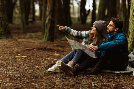 Positive young caucasian couple in jackets in forest enjoy journey, points finger at copy space, rest, look at mapの写真素材
