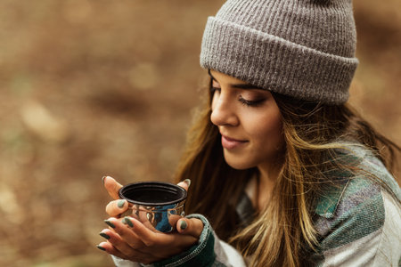 Cheerful young caucasian lady in jacket in forest drinks cup of tea, enjoy travel, vacation and hot coffeeの写真素材