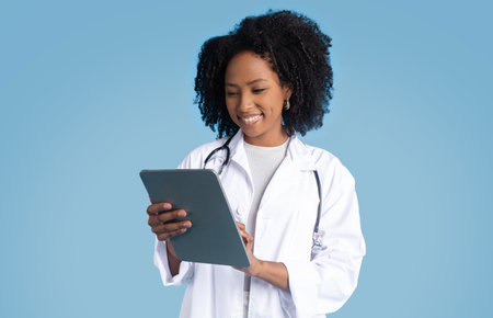 Happy black millennial lady doctor in white coat making notes in tablet, use app for chatの写真素材