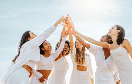 Carefree young multiracial women raising their beer bottles while standing on coastline at sunny day, having hen partyの写真素材