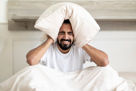 Annoyed Indian Guy Covering Ears With Pillow, Sitting On Bed At Homeの写真素材