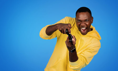 Happy excited millennial african american guy gamer in sweatshirt with open mouth and joystick...