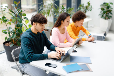 Three international classmates sitting at table in modern auditorium using various gadgets, preparing for examsの写真素材