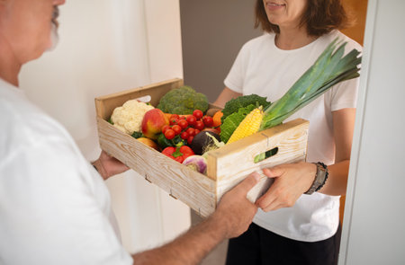 Serious old caucasian man taking box of organic vegetables from woman at home door, cropped, close upの写真素材
