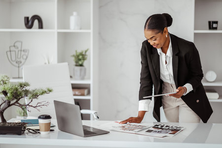 Positive black female editor checking magazine during working process satisfied with design, holding digital tabletの写真素材