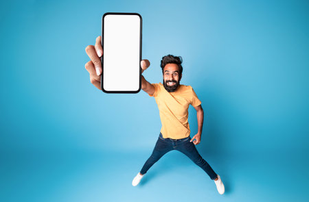 Happy indian man showing empty smartphone screen, jumping up over blue background, collage, mockup,...