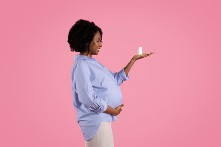 Cheerful young black pregnant woman touches big belly, enjoys motherhood, show jar of pills, vitamins and mineralsの写真素材