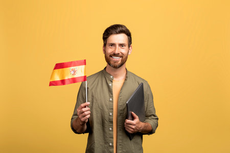 Cheerful middle aged man holding Spain flag and laptop, posing on yellow studio background and smiling at cameraの写真素材