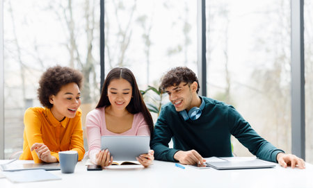 Happy diverse students using digital tablet, watching videos online sitting at table over window, copy spaceの写真素材