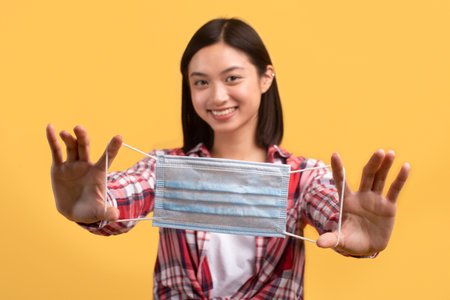 Virus protection concept. Happy asian lady holding medical face mask in hands and smiling, posing...