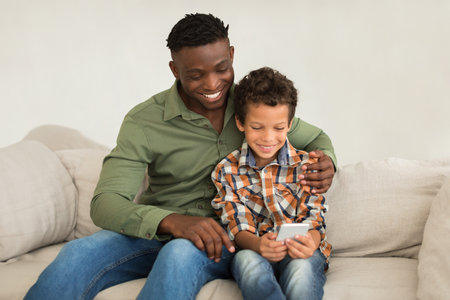 Smiling african american daddy and preteen son using cellphone indoorの写真素材