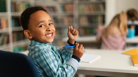 Positive black primary schoolboy sitting at desk in classroom, looking and smiling at camera, panorama, free spaceの写真素材