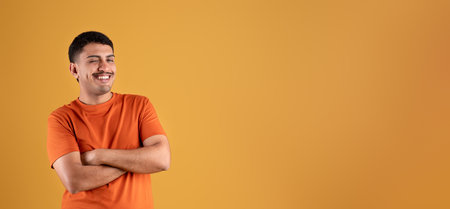 Positive brazilian man in orange t-shirt posing with crossed arms on chest, winking and smiling, panorama, free spaceの写真素材