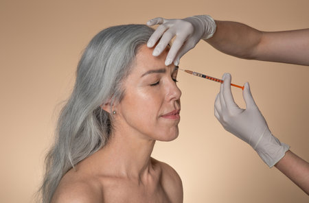 Senior woman with closed eyes on beauty procedure, cosmetologist making injection, isolated on beige backgroundの写真素材