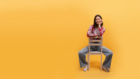 Smiling young chinese lady in casual sitting on chair isolated on yellow background, studio, panorama with free spaceの写真素材