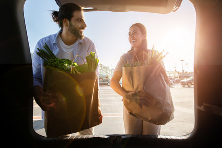 Young latin married couple putting paper bags full of healthy food into car on parking near mall or...