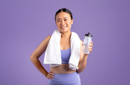 Excited asian woman in sportswear with towel holding bottle of water, enjoying sports on purple...