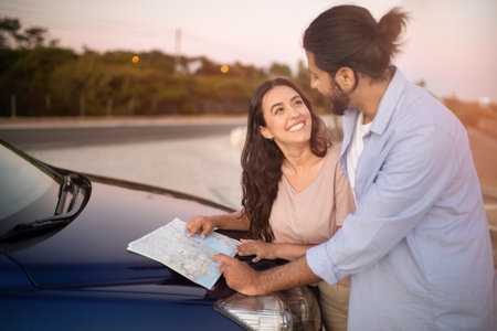 Enthusiastic latin couple reading map while having car trip, standing near the hood of car and...