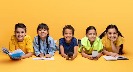 Cheerful multiracial children lying on floor, reading books, using phonesの写真素材