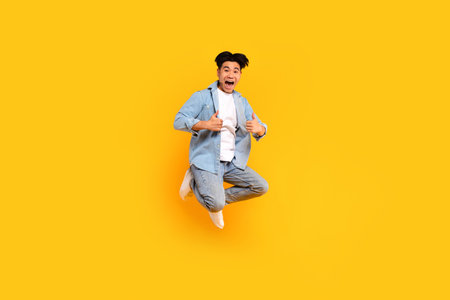 Excited asian man in stylish outfit showing thumb ups while jumping on yellow studio background,...