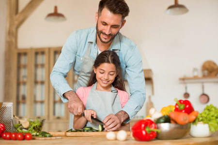Loving father teaching and helping his daughter to cook, cutting vegetables, preparing salad together in kitchenの写真素材