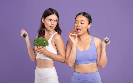 Asian woman eating donut while her friend holding broccoli and looking at her, standing over purple backgroundの写真素材