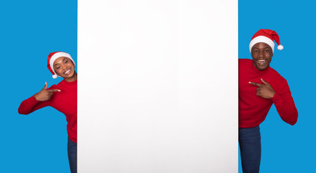 Black Couple In Santa Hats Pointing At Poster, Blue Backgroundの写真素材