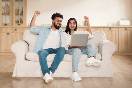 Giveaway, lottery, cashback. Overjoyed indian couple using laptop sitting on couch at home, gesturing and exclaimingの写真素材