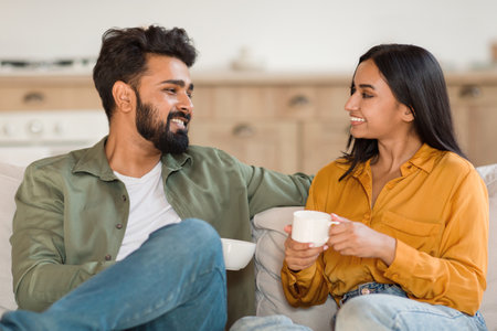 Weekend morning. Happy indian couple enjoying hot coffee and talking while sitting on sofa indoorsの写真素材